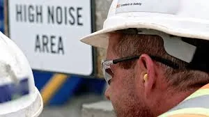 A worker wearing a hard hat and an inserted earplug standing in front of a HIGH NOISE AREA sign — the scenario OSHA's Hearing Conservation Program is written for
